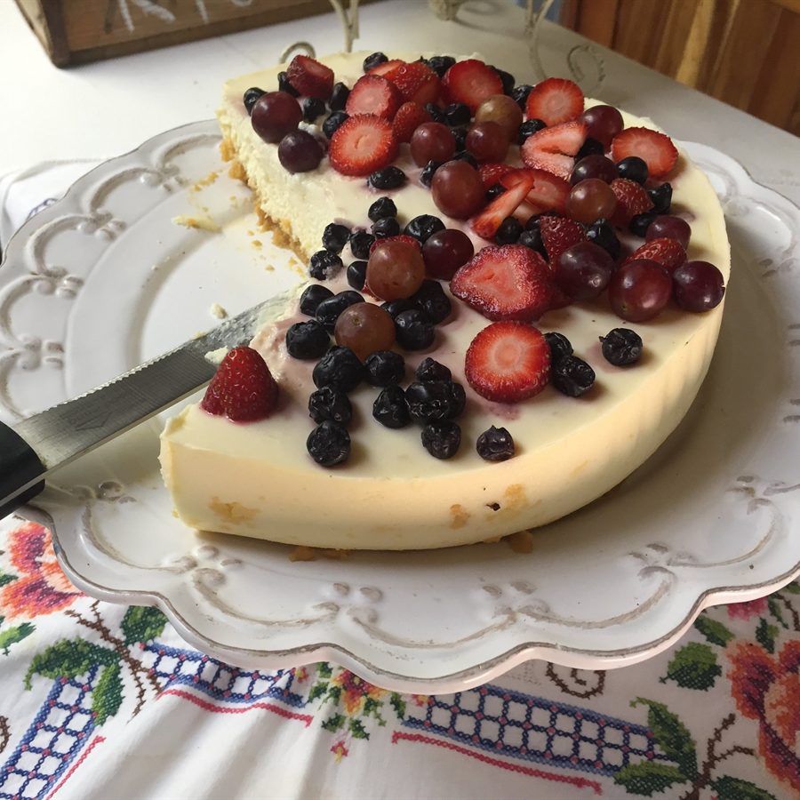 A whole cheesecake topped with strawberries, blueberries and grapes on a white cake stand.