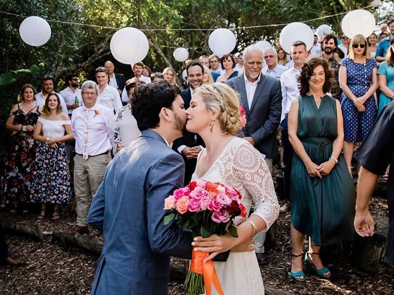 A couple kissing at their forest ceremony, surrounded by guests, with white paper lanterns hanging from the trees.